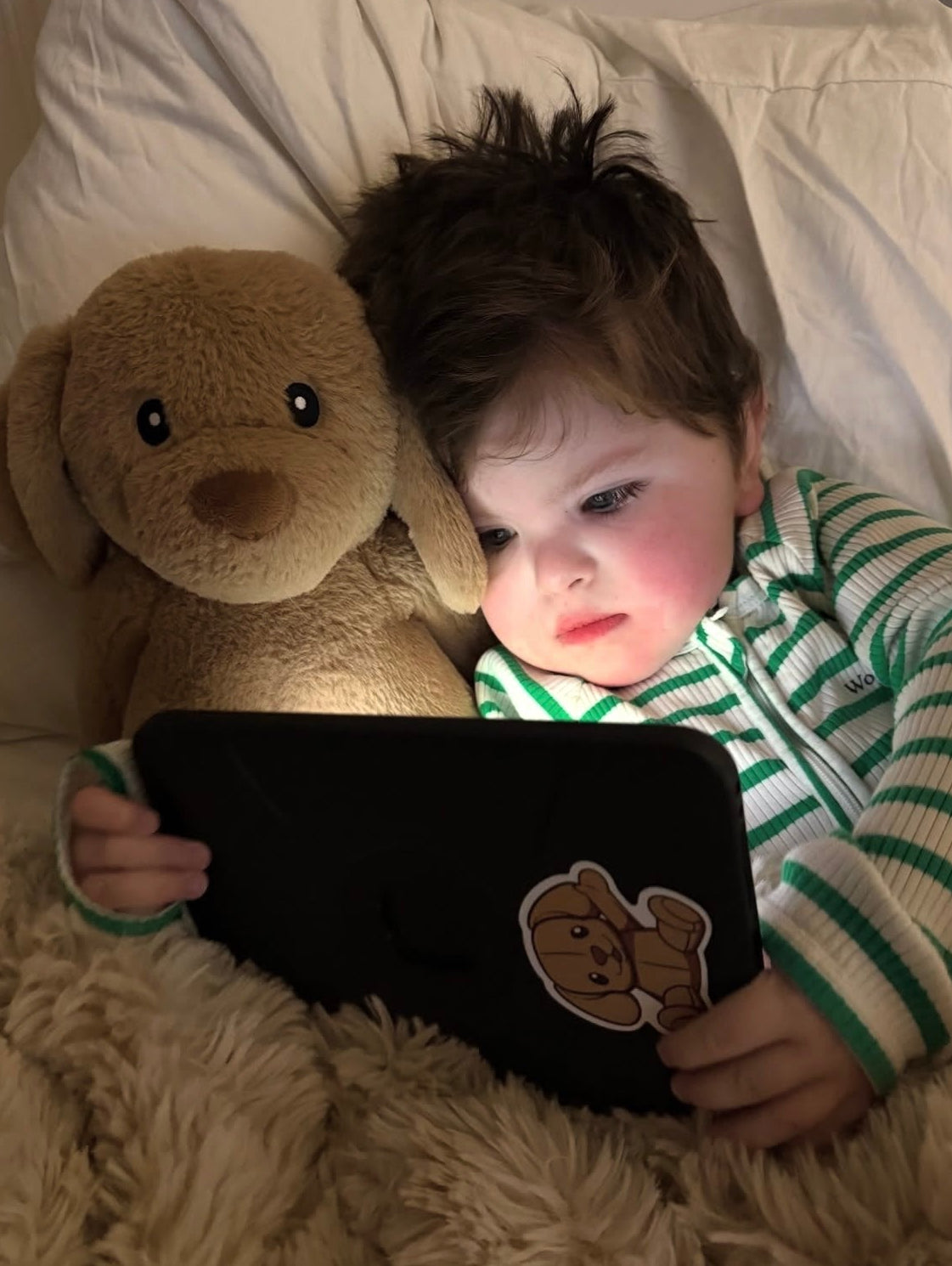 A toddler in a green and white striped pajama top lies on a fluffy cream blanket next to a brown plush dog toy, holding a tablet with a cartoon bear sticker.