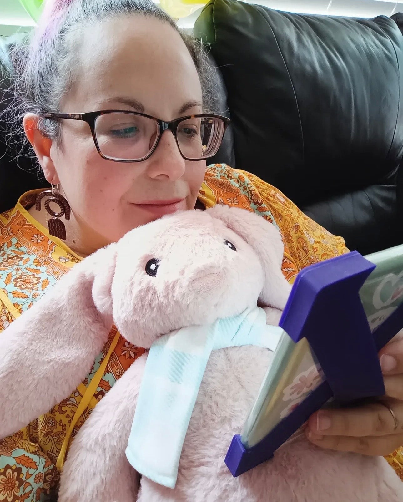 A woman with glasses and a pink hair streak is lying on a black leather couch, holding a light pink plush bunny with a light blue bow and looking at a purple tablet.